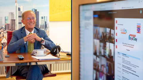 Staatsminister Wintermeyer sitzt am Schreibtisch und lächelt, neben ihm ein Monitor mit dem geöffneten Fenster der Live-Schalte.