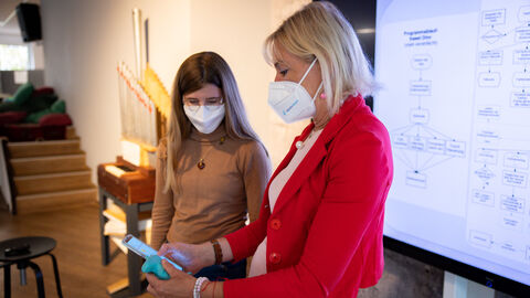 Lilly Scharz zeigt der Digitalministerin auf dem Handy ihre App. Beide tragen Mundschutz.