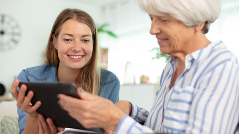 Eine junge Frau neben einer älteren Dame, die ein Tablet in der Hand hält. Beide lächeln und schauen auf das Tablet.