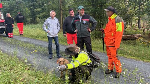 Mehrere Männer stehen zusammen. Einer der Männer zeigt einem Hund, wo er nach etwas suchen soll. 