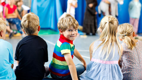 Kinder sitzen in eienr Reihe und gucken einer Theater Aufführung zu. Ein Kind dreht sich um und lacht in die Kamera.