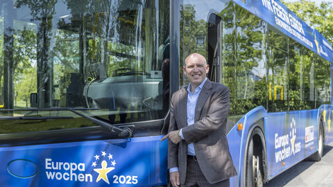 Manfred Pentz zeigt lachend auf einen blauen Europabus
