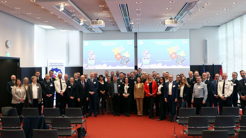 Eine Gruppe vieler Menschen steht zum Foto aufgereiht. Im Hintergrund sind Leinwände, auf denen eine Präsentation zu sehen ist.