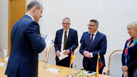 Zwei Männer stehen vor einem Tisch. Dahinter steht ein weiterer Mann, der zu ihnen spricht und ein Geschenk in der Hand hält.